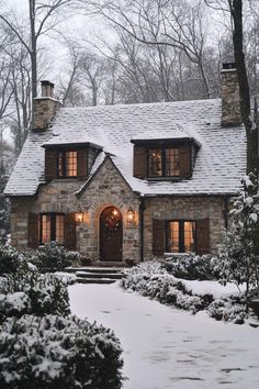 a stone house with lights on in the front yard and snow covered trees around it