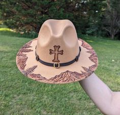 This unique fedora makes a great addition to any wardrobe. The warm tan material burns a rich deep brown allowing the design to stand out. A cross is intricately burned and shaded on the crown by hand using a pyrography machine. A detailed landscape of a mountain range and pine trees is sculpted and shaded along the entire circumference of the brim. All my hat designs are completely original and freehanded. For a beautiful finishing touch the hat is embellished with a western faux leather hat band.  Hat Materials: Cotton and polyester Hat fits most women with an adjustable tie band inside *Disclaimer* Colors may vary slightly screen to screen Detailed Landscape, Antler Hat, Cowboy Hat Design, Hat Burning, Custom Cowboy Hats, Accessories Boho, Painted Hats, Hat Fits, Leather Hat