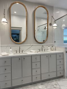 two oval mirrors are above the double sinks in this bathroom with gray cabinets and marble countertops