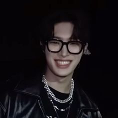 a young man wearing glasses and a black leather jacket smiles at the camera in front of a dark background