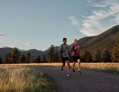 two people running down a dirt road in the middle of a field with mountains behind them