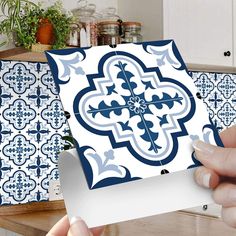 a person holding up a piece of paper with blue and white designs on it in front of a kitchen counter