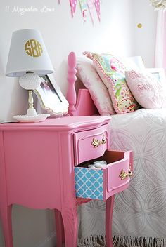 a pink dresser with two drawers and a lamp on top of it next to a bed