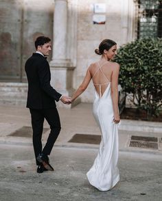 a bride and groom hold hands as they walk down the street in front of an old building