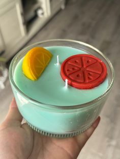 a hand holding a small glass container filled with fruit and candle wax on top of a wooden floor