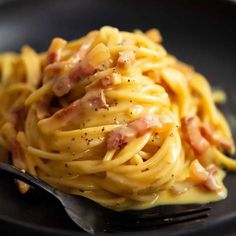 a black plate topped with pasta covered in ham and cheese sauce next to a fork