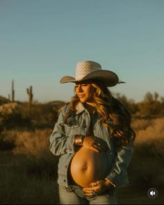 a pregnant woman wearing a cowboy hat and jeans is holding her belly in the desert