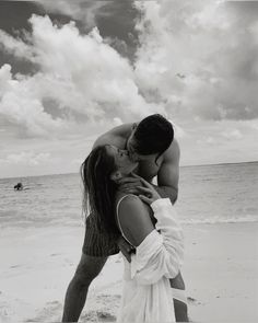 a man kissing a woman on the beach with clouds in the sky behind them and people swimming in the water