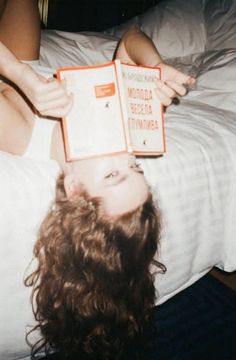 a woman laying in bed with her head on top of a book that is upside down