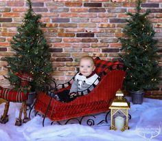 a baby sitting in a sleigh next to christmas trees and a brick wall