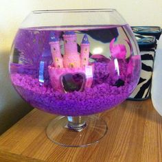 a fish bowl filled with purple sand and pink castle in the middle on top of a wooden table