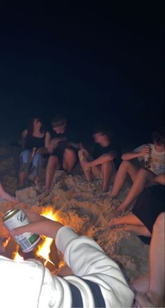 a group of people sitting around a campfire on the beach with drinks in their hands