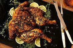 a chicken with lemons and herbs on a black plate next to a knife and fork