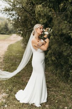 a woman in a white wedding dress holding flowers and a veil over her head while standing on the grass