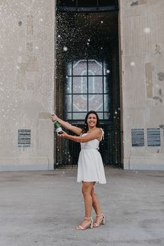 a woman standing in front of a building holding a bottle and throwing snow into the air