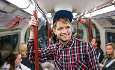 a man in plaid shirt and hat on train with other people sitting behind him looking at the camera