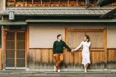 a man and woman holding hands while standing in front of an old building with bamboo shutters