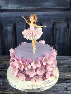 a birthday cake decorated with pink flowers and a ballerina figure on top, sitting on a wooden table