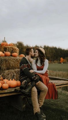 a man and woman sitting on a wooden bench next to hay bales filled with pumpkins