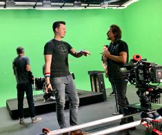 two men standing in front of a green screen with camera equipment on the floor and behind them