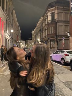 two women are hugging on the street in front of parked cars and buildings at night