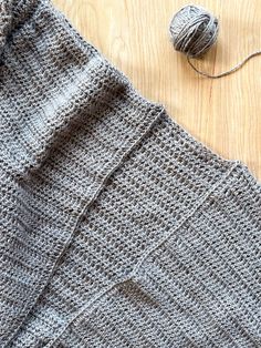 a gray knitted sweater next to a ball of yarn on a wooden table with a crochet hook