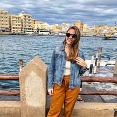 a woman standing in front of a body of water wearing brown pants and a denim jacket