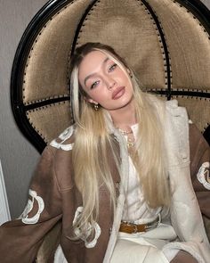 a woman with long blonde hair sitting in a chair wearing a brown and white jacket