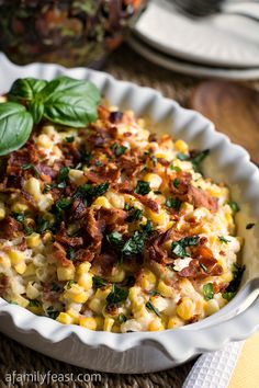 a white bowl filled with corn and bacon on top of a table next to silverware