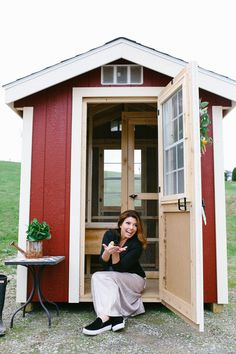 a woman sitting in the doorway of a small red and white chicken coop with her legs crossed