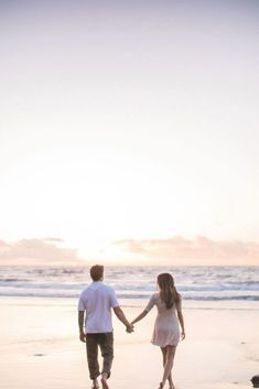 a man and woman holding hands while walking on the beach at sunset with waves coming in