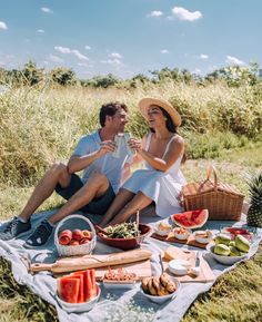 a man and woman sitting on a blanket with food in front of them, smiling at each other