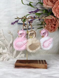 three little pink and white birds hanging from a wooden stand with flowers in the background