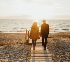 a man and woman walking down a wooden walkway towards the ocean on a sunny day