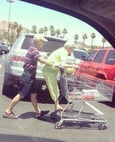 an older man pushing a shopping cart through a parking lot next to a parked car