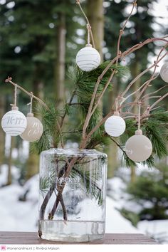 a glass vase filled with ornaments on top of a wooden table next to trees in the snow