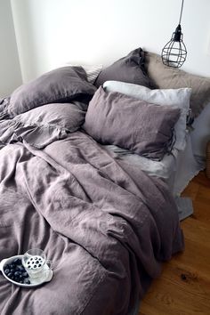 an unmade bed with grey linens and pillows on the floor next to a lamp