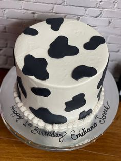 a birthday cake decorated with black and white cow spots on a wooden table next to a brick wall