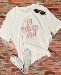 a white shirt with the words pi beta phi on it next to sunglasses and a pair of sneakers