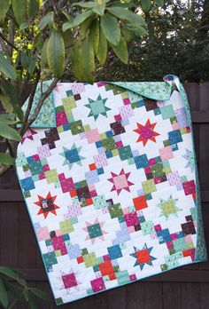 a quilt hanging from a tree in front of a fence with green leaves on it