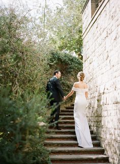 a bride and groom walking up some steps together in front of a brick wall holding hands