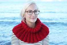 a blonde woman wearing glasses and a red knitted cowl neck scarf by the ocean