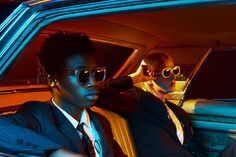 two men sitting in the back seat of a car, one wearing sunglasses and the other with dark hair