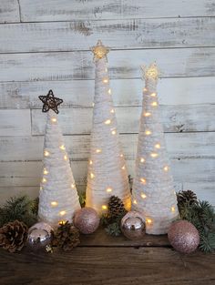 three white christmas trees decorated with lights and pine cones on top of a wooden table