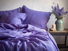 a bed with purple sheets and pillows next to a vase with lavender flowers on it