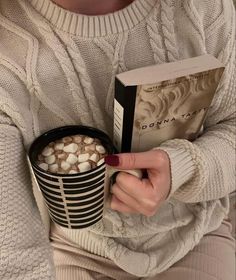 a woman holding a cup of hot chocolate and a book in her hands while sitting on a couch