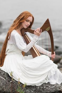 a woman in white dress holding a harp on top of a rock next to the ocean