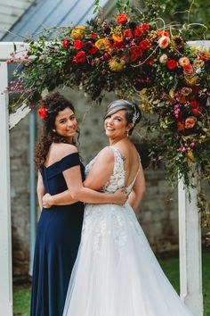 two women standing next to each other under a floral arch