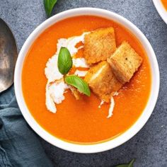 two bowls of tomato soup with croutons and basil on the side next to a spoon