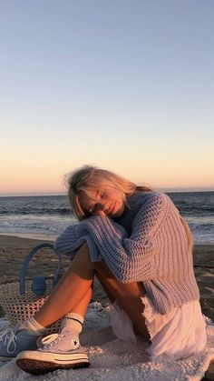 a woman sitting on top of a sandy beach next to the ocean with her legs crossed